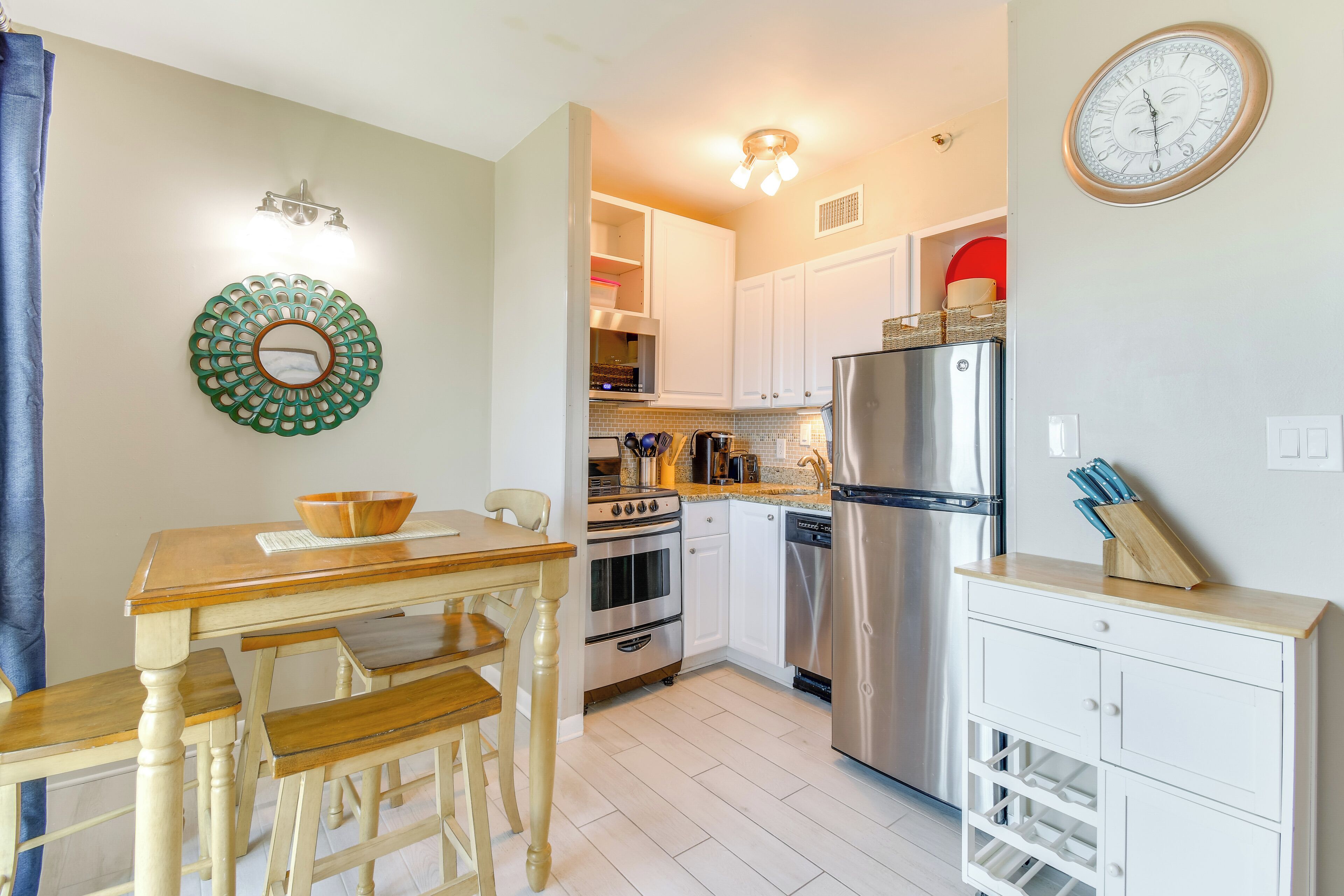 A compact kitchen with white cabinets, a stainless steel refrigerator, a wooden dining table with benches, and a wall-mounted clock.