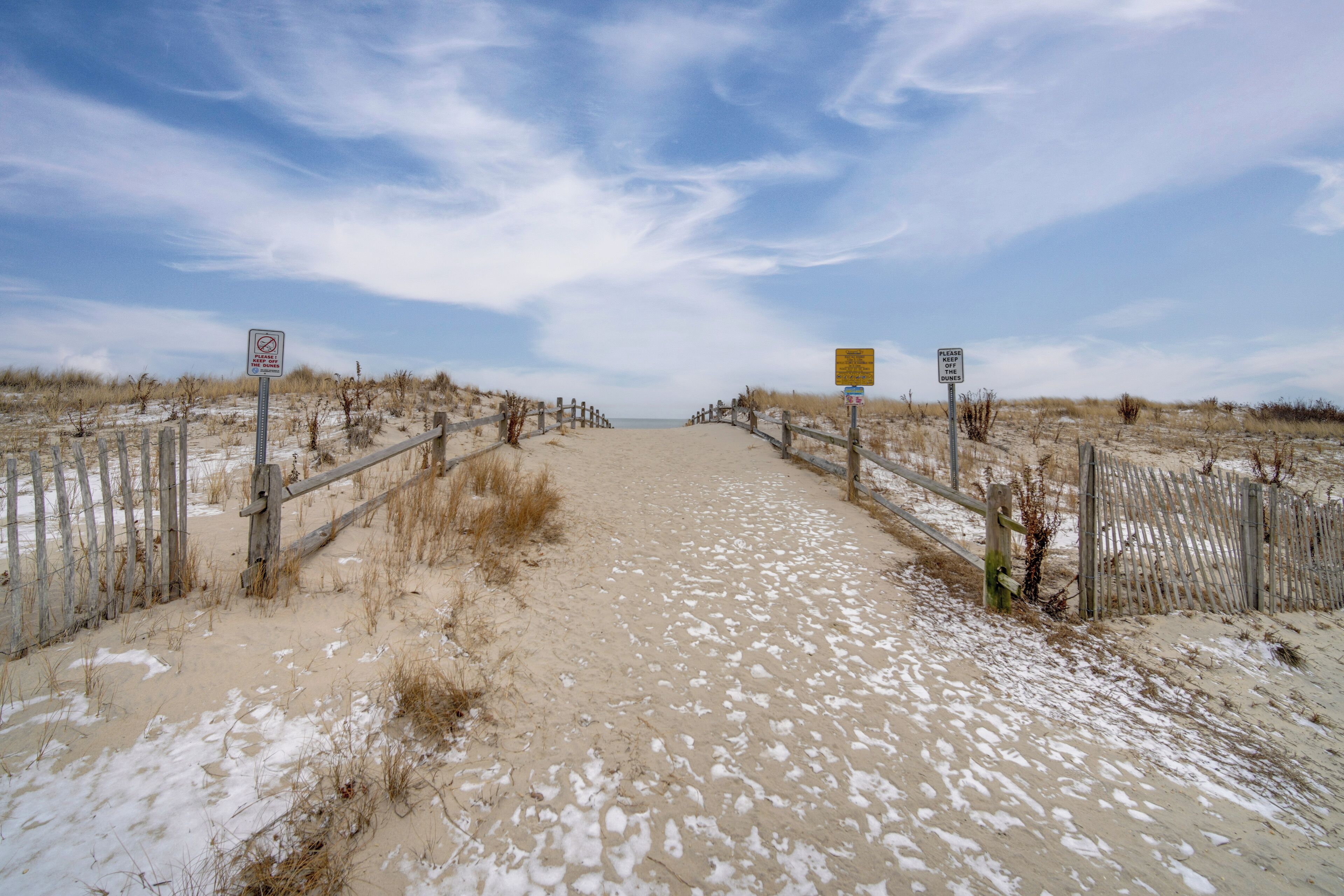 A sandy beach path with wooden fences and signs, leading to the ocean.