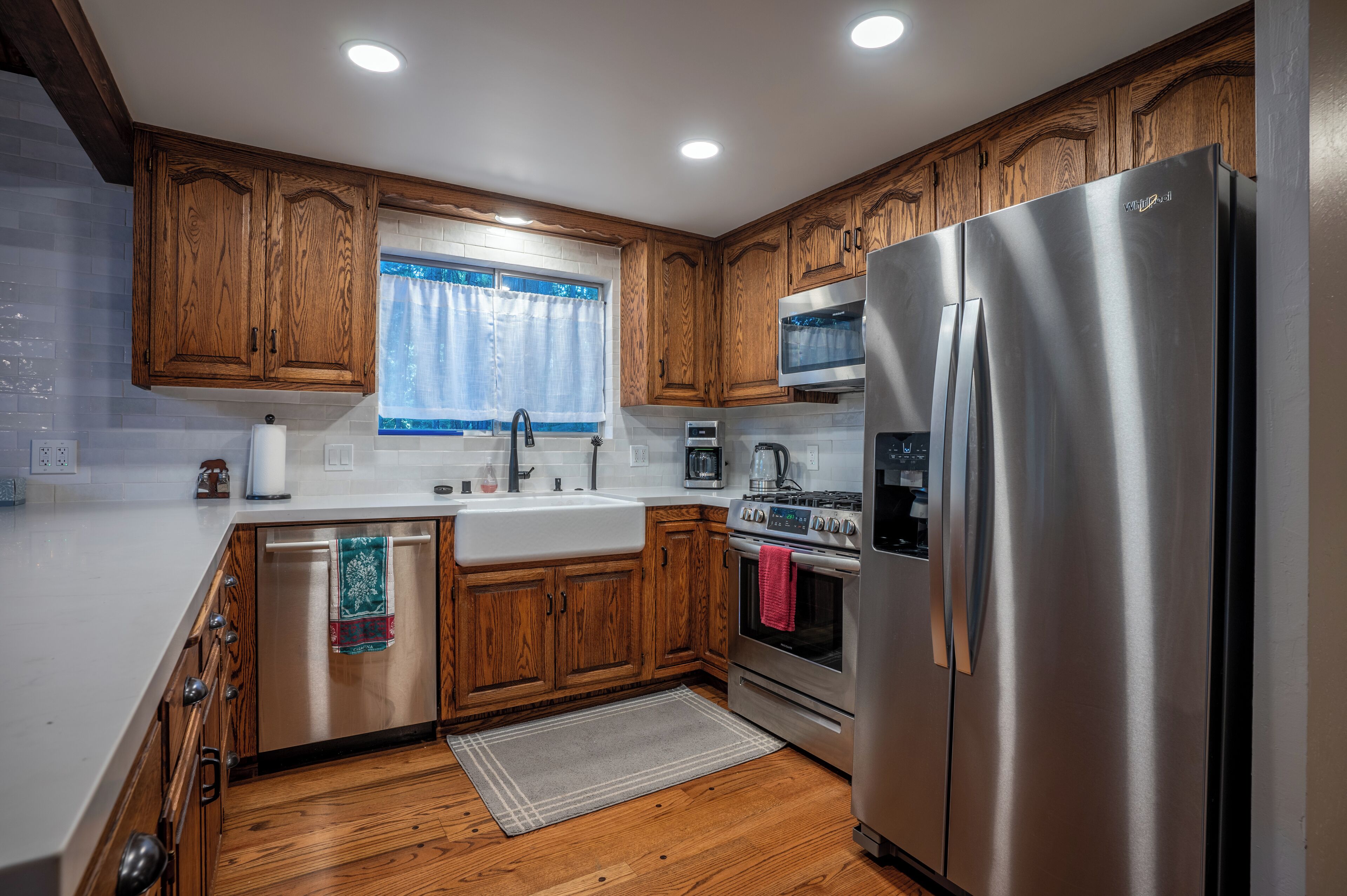 A kitchen with wooden cabinets, stainless steel appliances, a window with curtains, and a tiled backsplash.