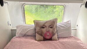 A bed with a pink quilt, two pillows, and a window with a view of greenery.