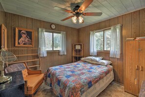A bedroom with wooden walls, a bed with a colorful quilt, a ceiling fan, a desk with a lamp, and a window with curtains.