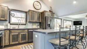 A kitchen with wooden cabinets, a large island with bar stools, a clock, and a refrigerator. - Boat Dock: Riverfront Oconto Home! (Oconto)