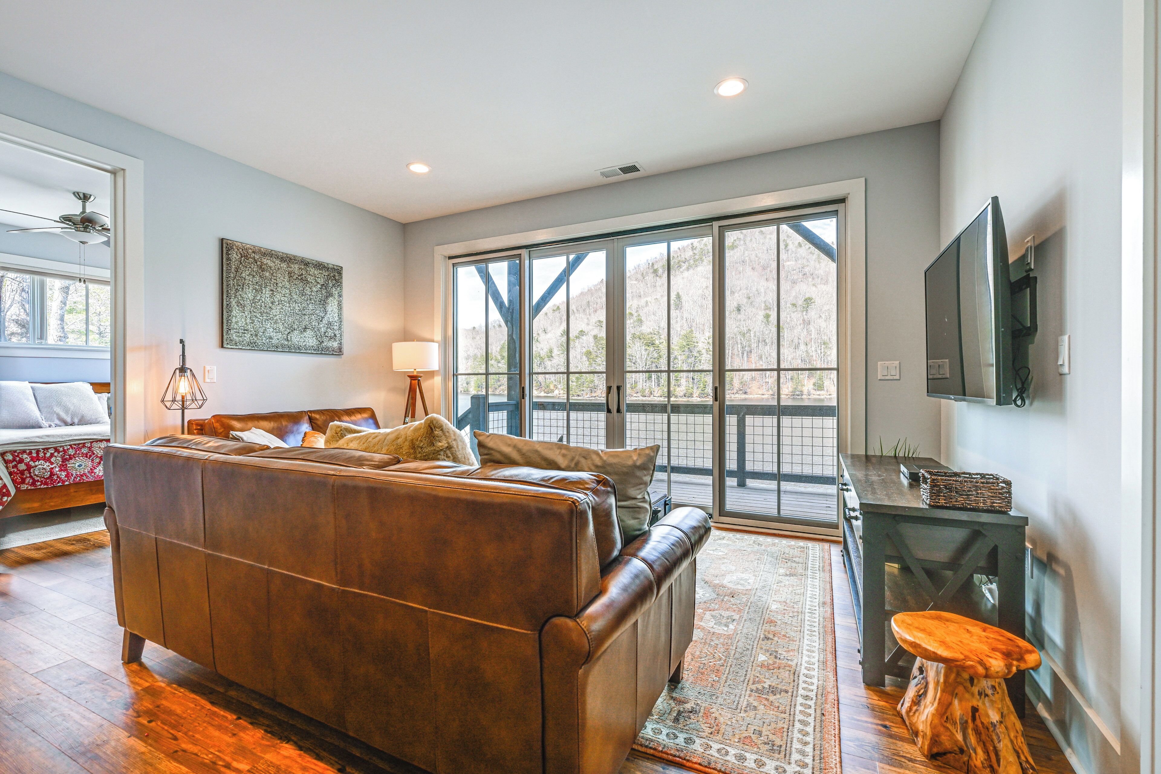 A spacious living room with a brown leather sofa, a wooden coffee table, and a large window offering a view of trees.