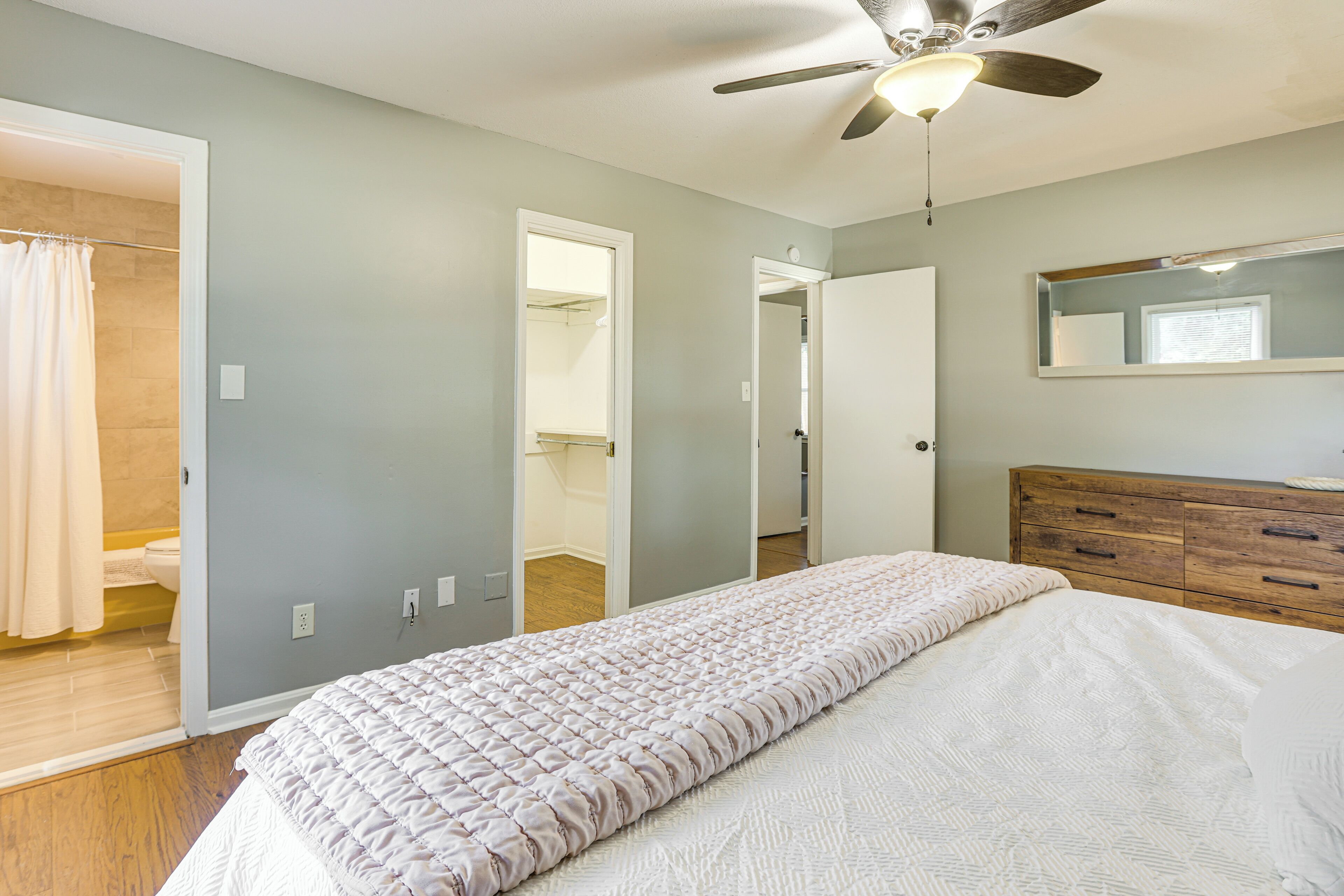 A bedroom with a bed, a wooden dresser, a ceiling fan, and a mirror on the wall.