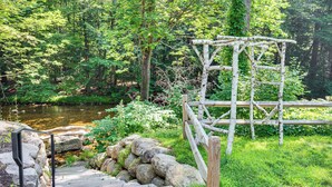 A wooden deck leading to a rustic wooden structure near a river, surrounded by lush greenery. - Walkable North Woodstock Home on Main Street! (North Woodstock)