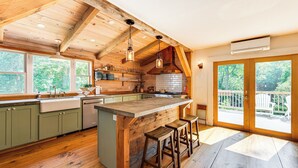 A spacious kitchen with wooden beams, a large island with stools, and a view of the outdoors through large windows. - Walkable North Woodstock Home on Main Street! (North Woodstock)