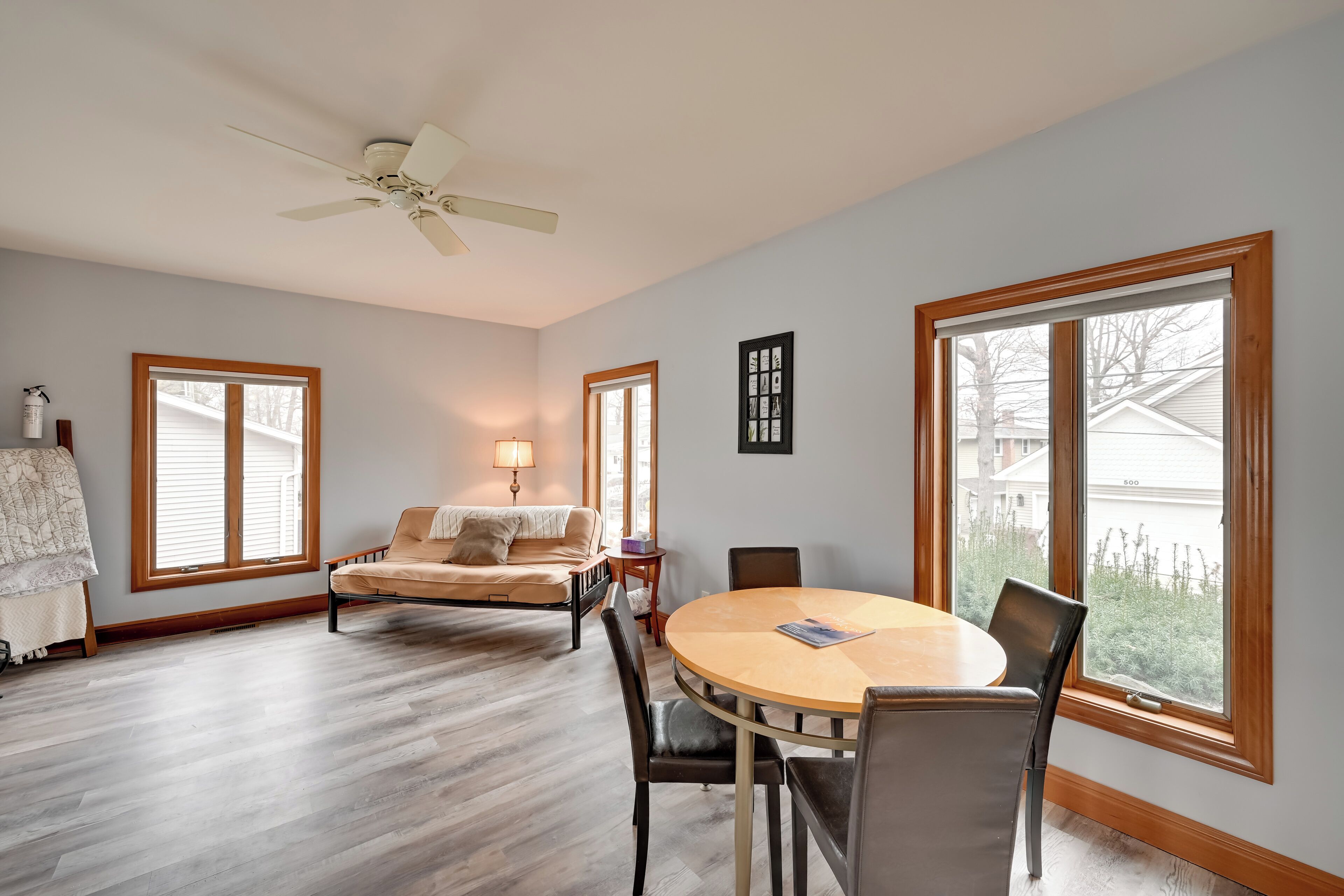 A spacious living room with hardwood flooring, a round wooden table with chairs, a beige sofa, and a ceiling fan. Large windows provide natural light and views of the outside.