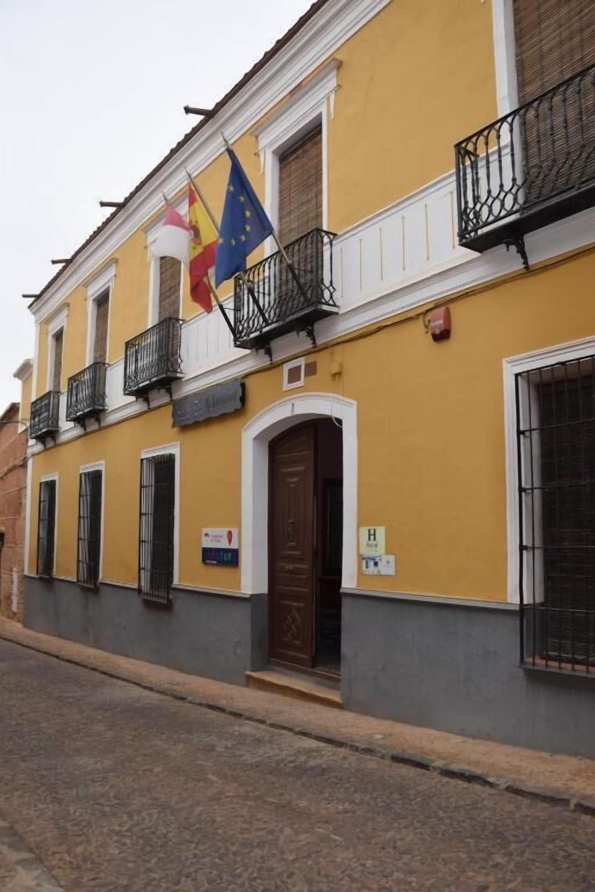 Un edificio amarillo con puerta marrón, balcones negros y banderas, incluyendo la bandera de la UE.