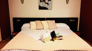 Ein Hotelzimmer mit einem großen Bett, mehreren Kissen, einem Nachttisch mit Buch und Flasche sowie einem Bild an der Wand.