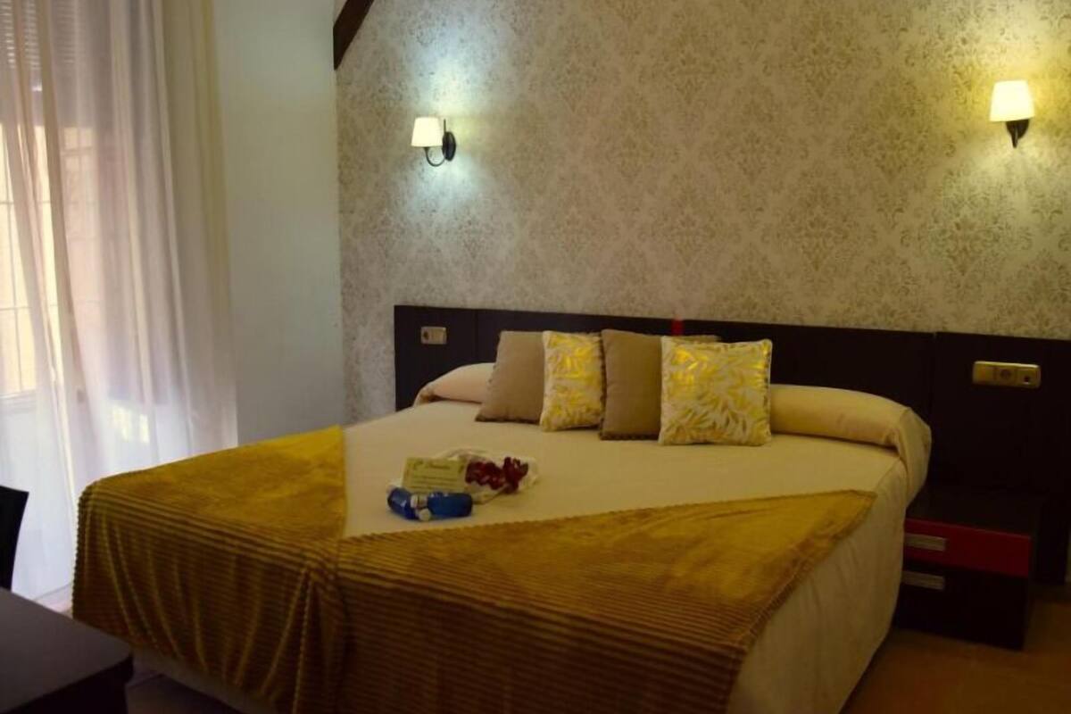 Une chambre d’hôtel avec un grand lit, deux lampes fixées au mur, une fenêtre avec des rideaux et une table de chevet.