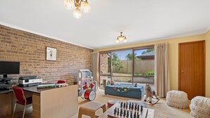 A cozy living room with a brick wall, a television, a desk with a computer, a drum set, a chess table, and a rocking horse. - Spacious Family Retreat on Fischer (3) (Torquay)