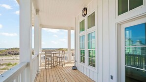 A wooden deck with a white railing, a table with chairs, and a view of the beach and ocean.
