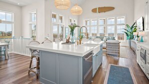 A spacious kitchen with a large island, white cabinetry, and a view of the ocean. There are wicker chairs and a dining table, and the room has large windows and pendant lights.