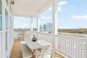 A covered outdoor area with a wooden deck, white railings, and a view of a large blue house across the beach.