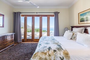 A bedroom with a large bed, a dresser, a ceiling fan, and a view of the ocean through a sliding glass door. - Camps Bay 4bed Home w Pool and Views Alma Marina (Cape Town)