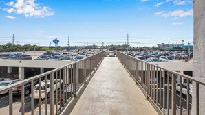 A pedestrian bridge leading to a parking area with multiple cars, a clear sky, and distant buildings.