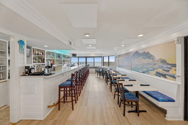A long dining area with high tables and chairs, a bar on the left, and a large window with a cityscape view.