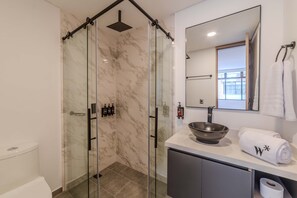A modern bathroom with a glass-enclosed shower, a marble-tiled wall, a white sink with a large bowl, and a towel with a monogram.