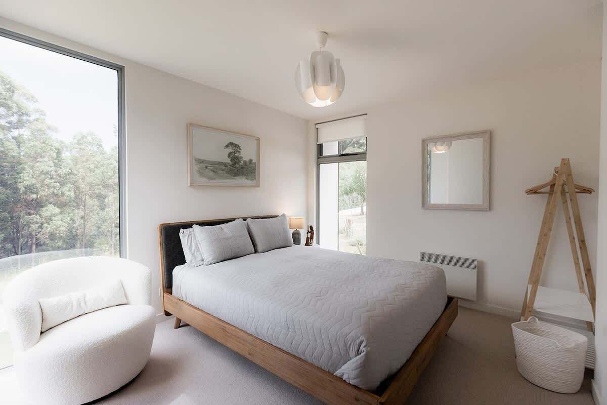 Une chambre à coucher moderne avec un grand lit, un fauteuil blanc, une lampe sur pied et une vue sur l’extérieur.