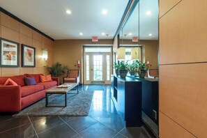 Un hall con un sofá rojo, una mesa de centro, un mostrador de recepción con plantas en macetas y una puerta que da al exterior.