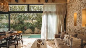 A modern living room with a pool view, wooden furniture, and a stone wall.