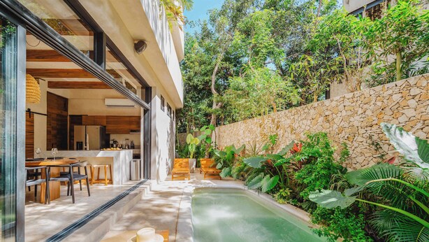 A modern outdoor dining area with a pool, stone wall, and lush greenery.