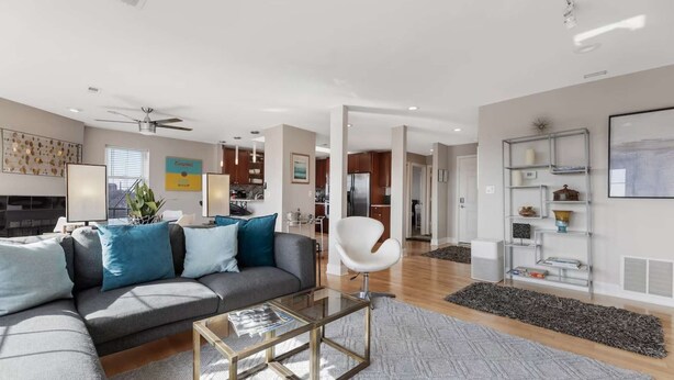 A modern living room with a sofa, coffee table, and a white chair. There is a kitchen in the background with wooden cabinets and a refrigerator. - Penthouse Style Condo with City View (Cincinnati)