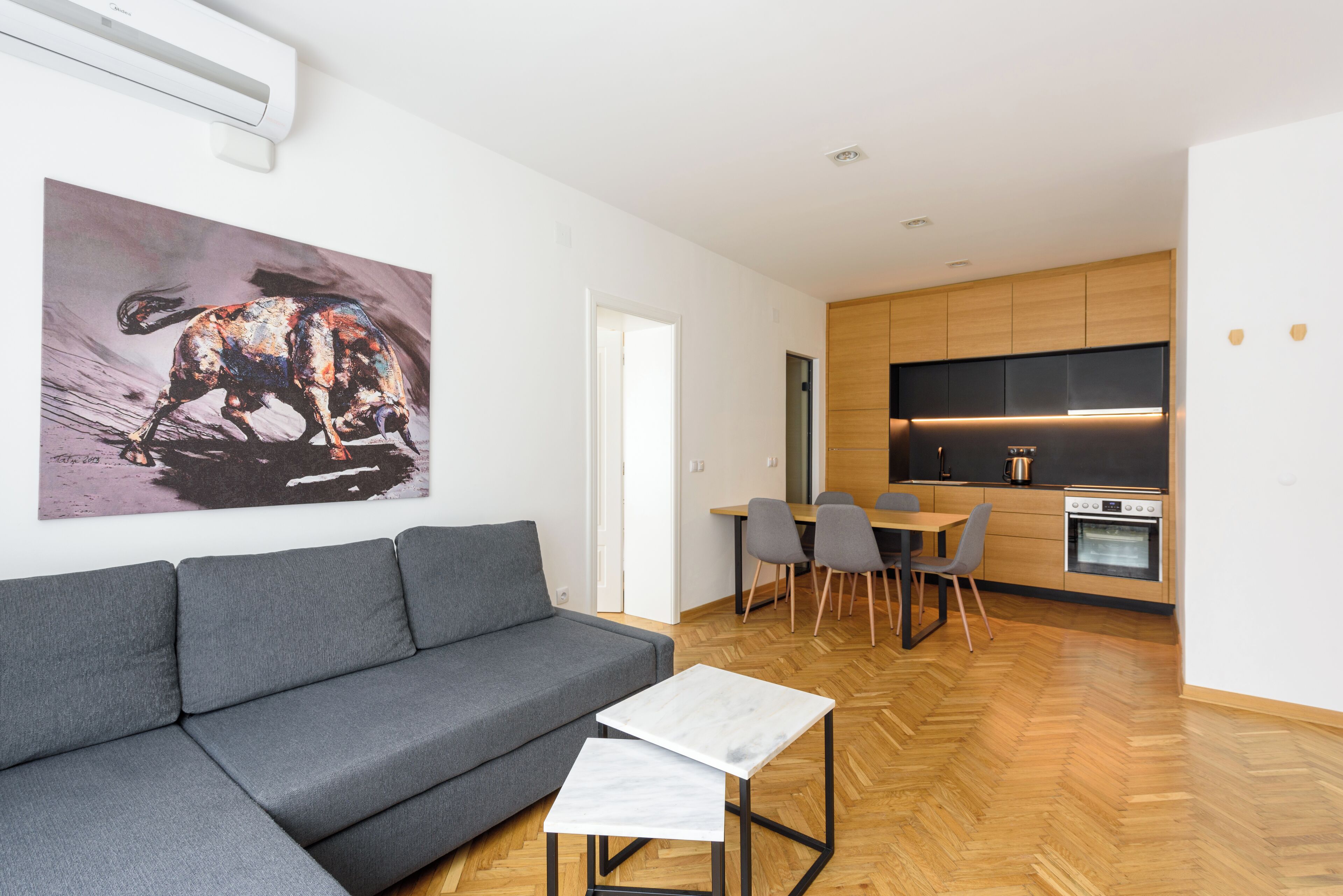 A modern living room with a grey sofa, a small white table, and a dining area with chairs and a table. There is a kitchen with wooden cabinets and a built-in oven. A large abstract painting is on the wall.
