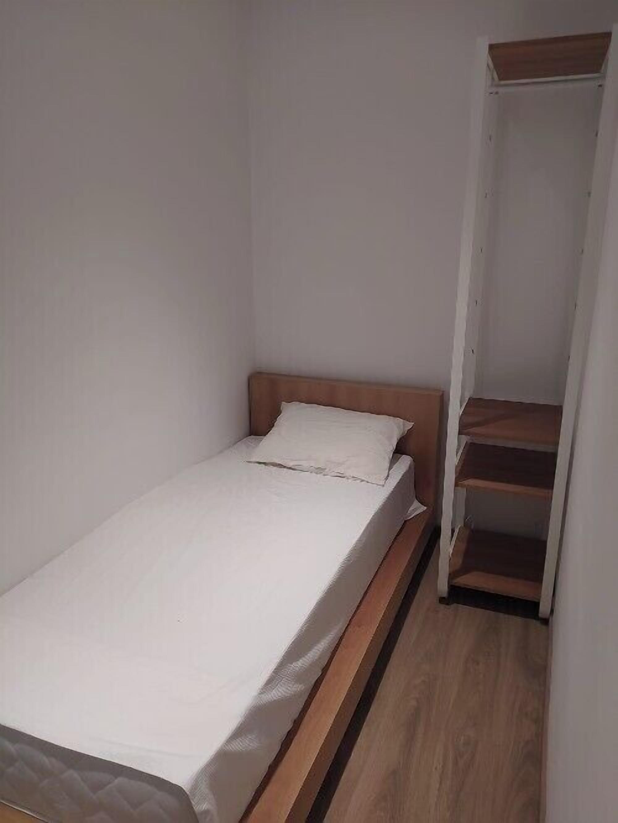 Une petite chambre avec un lit simple, une tête de lit en bois et un matelas blanc. On y trouve une armoire en bois avec un miroir et une étagère sur le côté droit.