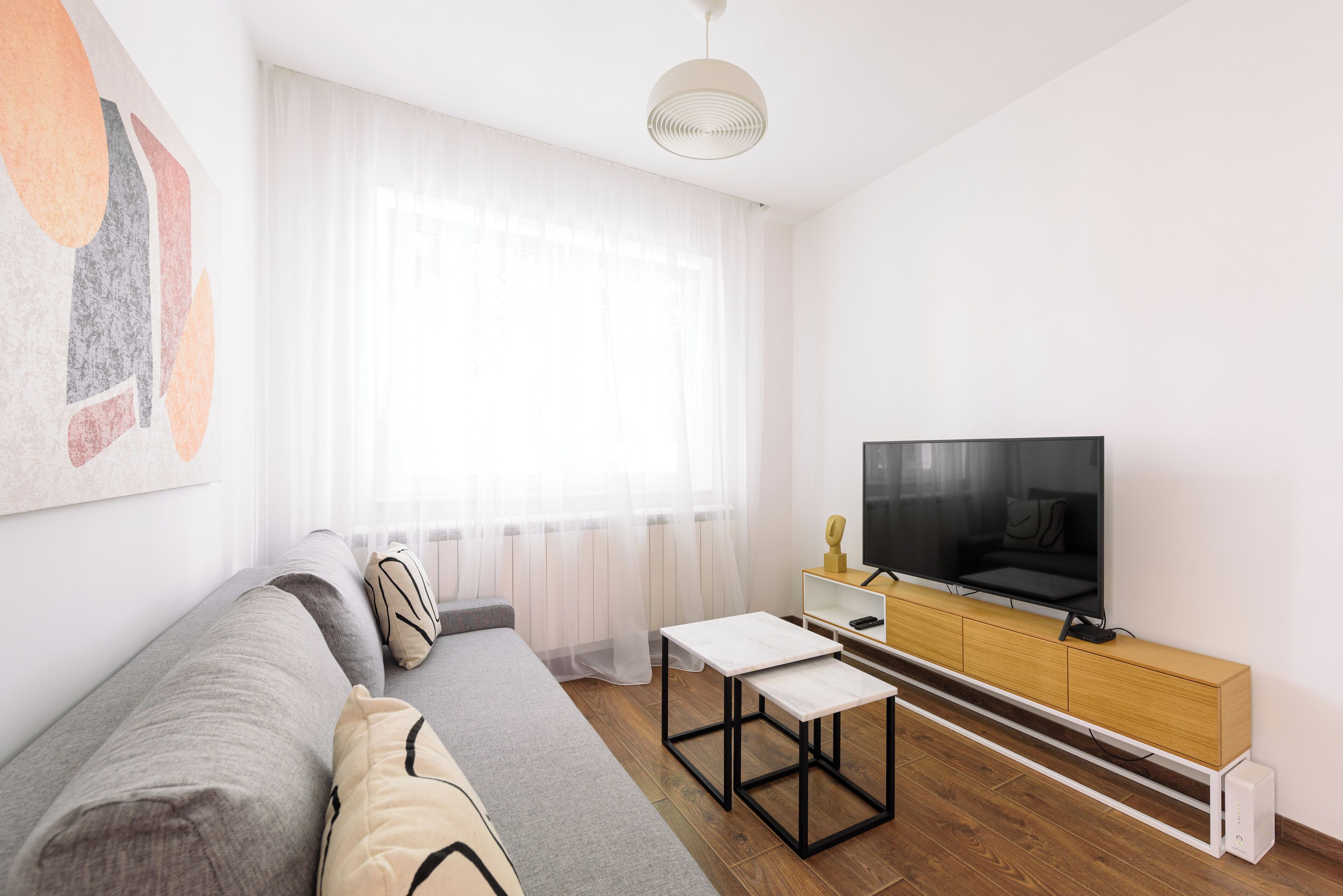 A modern living room with a sofa, a coffee table, a TV on a wooden cabinet, and a large window with sheer curtains.