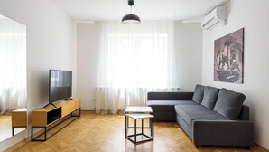 A modern living room with a wooden herringbone floor, a grey sofa, a TV on a wooden stand, and a pendant light. - Tim 3 (Belgrade)