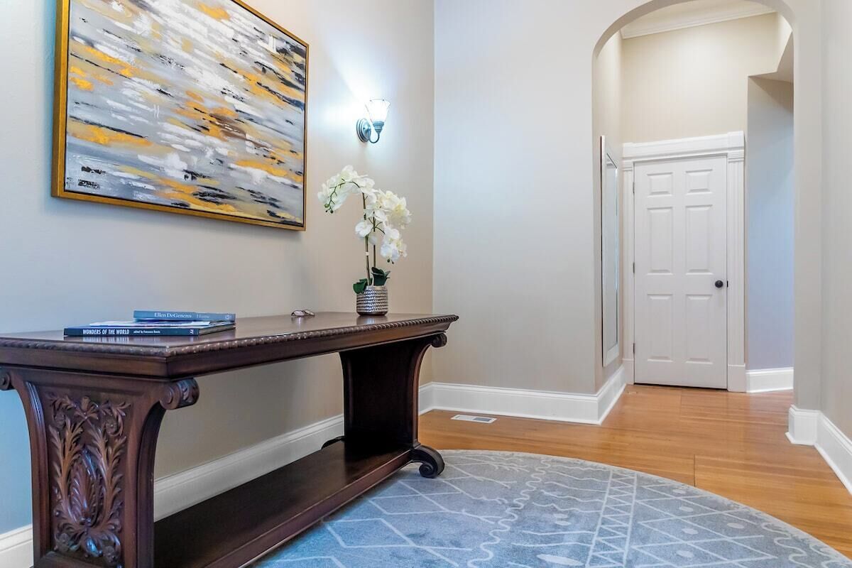 A hallway with a dark wooden console table, a white orchid in a vase, a large abstract painting, and a white door at the end.