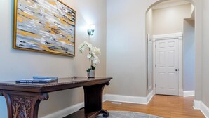 Un pasillo con un mueble consola de madera oscura, una orquídea blanca en un jarrón, un cuadro abstracto grande y una puerta blanca al final.
