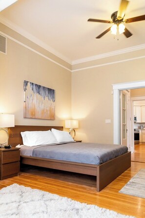 A bedroom with a bed, two bedside lamps, a ceiling fan, a painting on the wall, and a view into the kitchen.