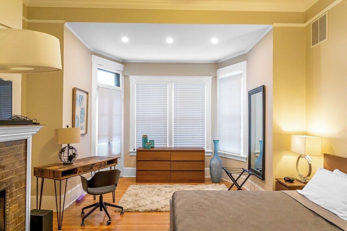 A bedroom with a bed, desk, chair, dresser, and a fireplace.
