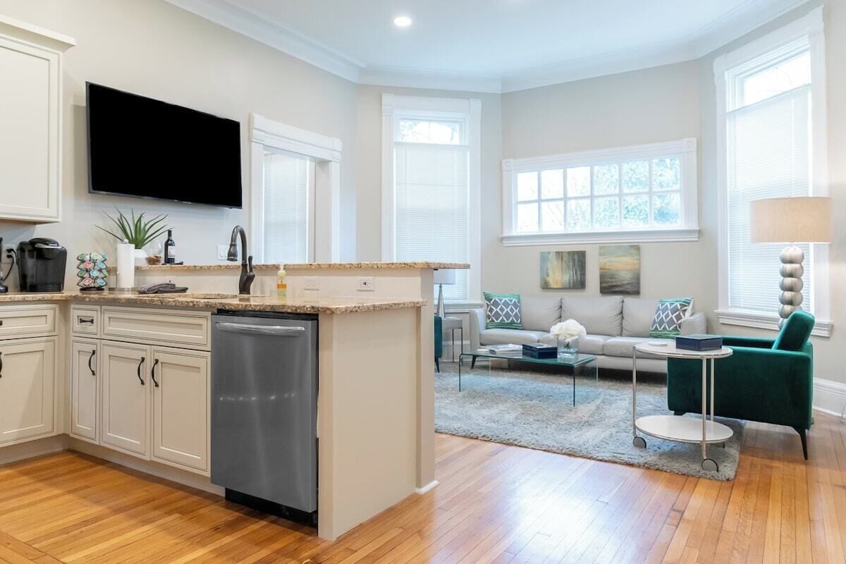 A modern kitchen with a dishwasher, a flat-screen TV, and a dining area with a green chair and a round table.