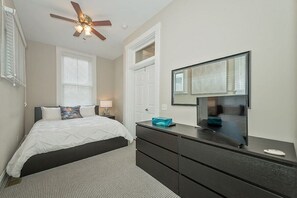 Une chambre à coucher comprenant un lit, une commode, un téléviseur, un ventilateur de plafond et une fenêtre avec des stores.