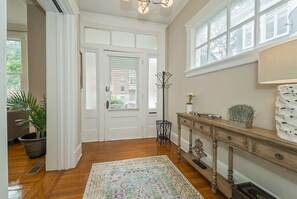 Une pièce avec un plancher en bois, un tapis à motifs, une porte blanche, une fenêtre, une console ornée d’objets d écoratifs et une plante en pot.