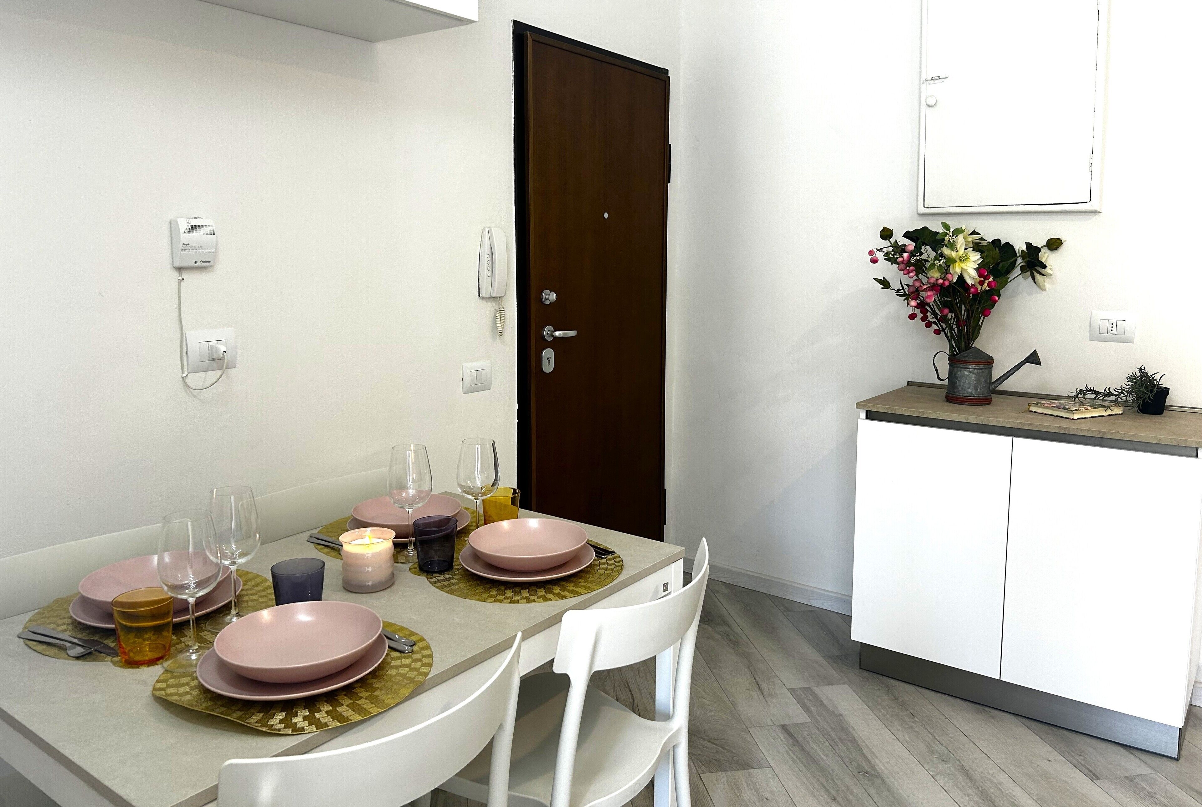 A dining area with a table set for four, a white chair, a cabinet, and a vase with flowers.