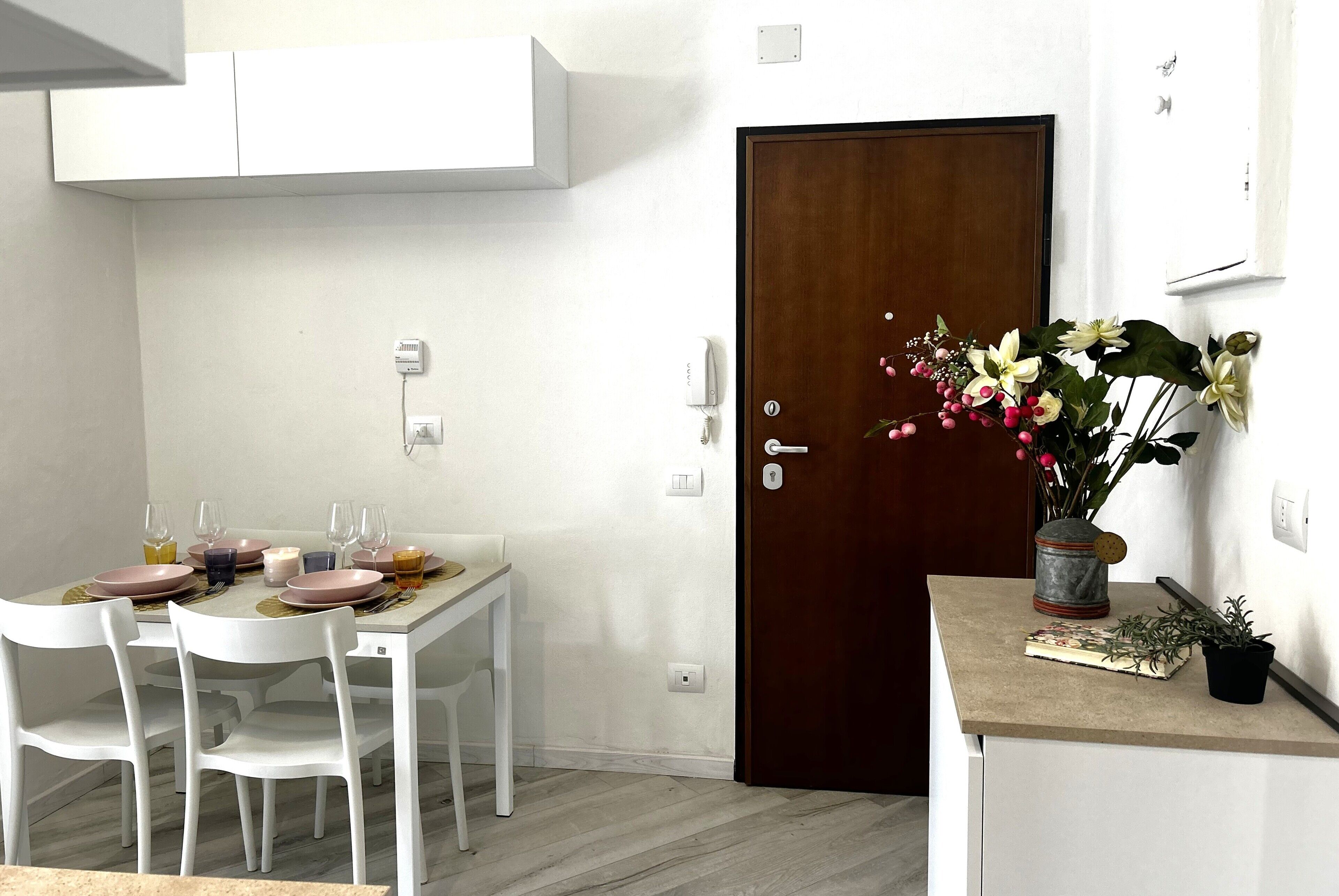 A dining area with a table set for four, white chairs, a wooden door, and a small potted plant on a counter.