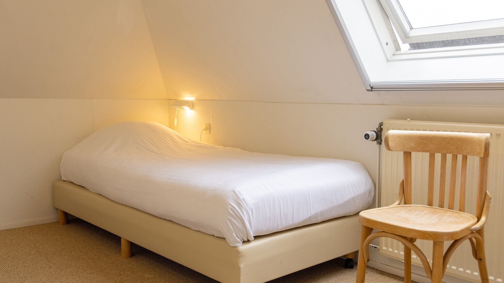 Une petite chambre avec un lit simple, un plafond en pente et un puits de lumière. On y trouve une chaise en bois et un radiateur fixé au mur.