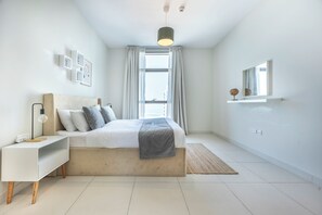 A modern bedroom with a bed, bedside table, lamp, and a view of the city through the window. - Bloomfields Premium 1BR Reflection (Abu Dhabi)