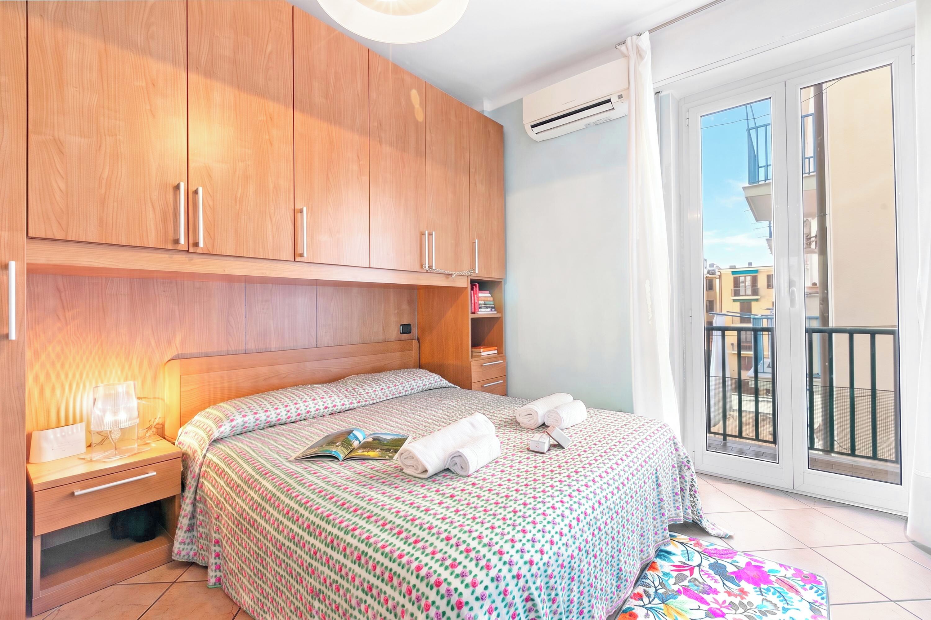 A bedroom with a wooden wardrobe, a bedside table with a lamp, a bed with a floral bedspread, and a view of the outside balcony.