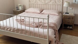 A bedroom with a white metal bed frame, a wooden nightstand with a lamp, and a bedside table with a book and a small plant. - Melloni Street (Dolo)