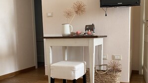 A small, neatly arranged room with a white table, a white cushioned stool, a white vase with dried flowers, a small house-shaped container, and a television mounted on the wall. - Melloni Street (Dolo)