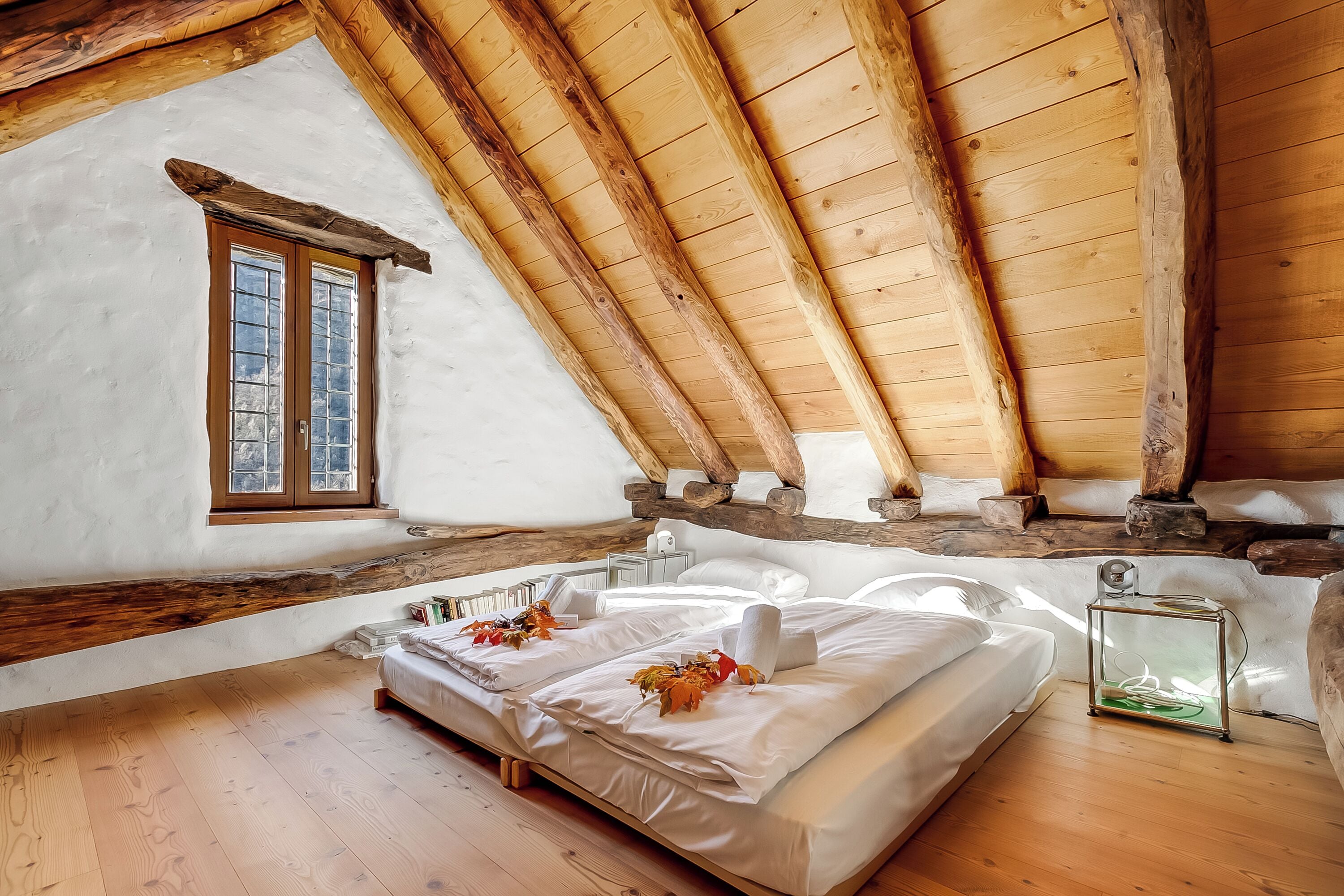 Un dormitorio acogedor en el ático, con techo de madera, un ventanal grande y una cama con sábanas blancas y hojas de otoño.
