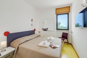 A neatly made bed with a beige bedspread, a red chair, a small table with a lamp, and a window with a view of greenery. - La Rosa Apartment Sea View (Salve)