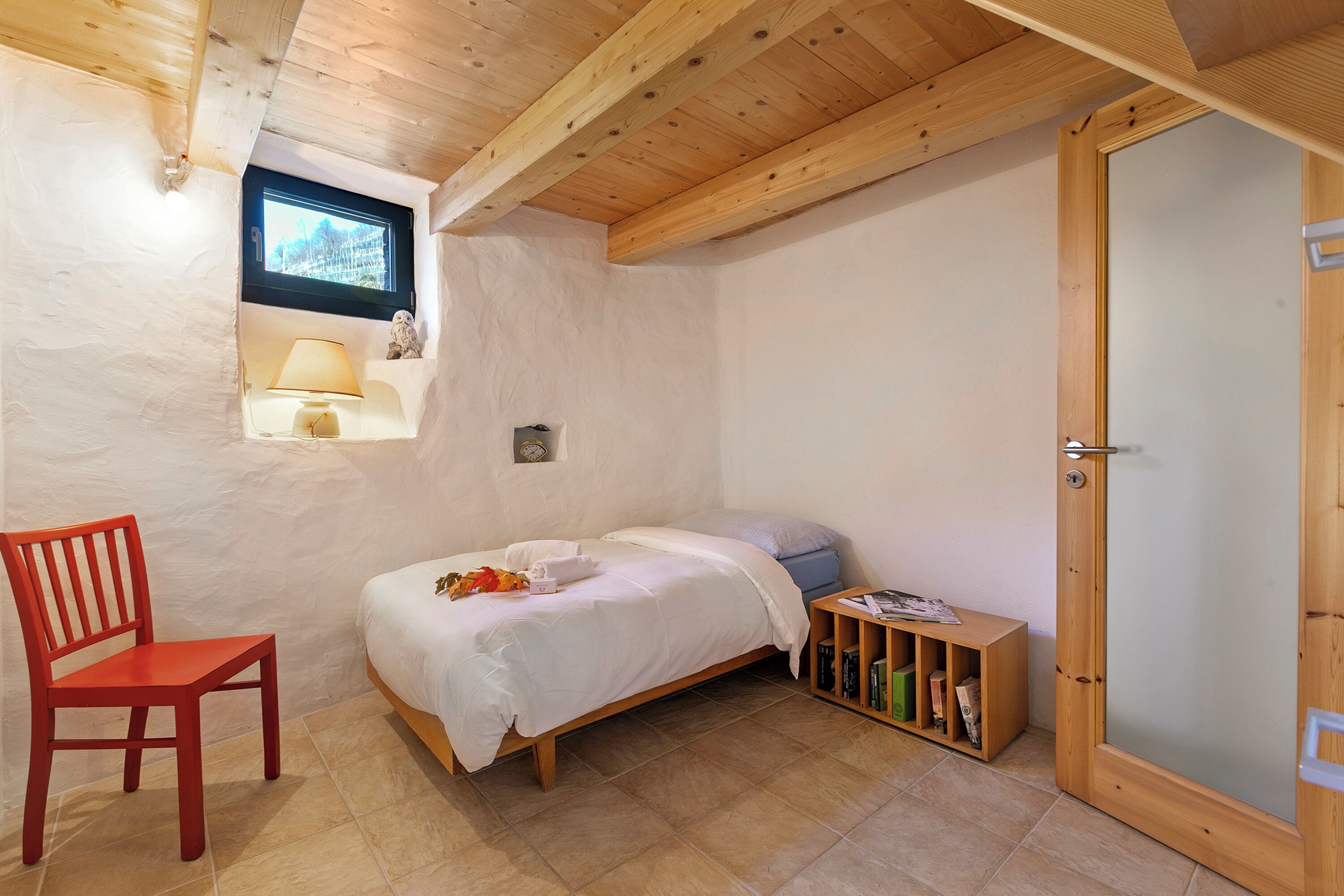 Une chambre avec un plafond en bois, un lit avec une literie blanche, une chaise rouge, une petite fenêtre et une porte en bois.