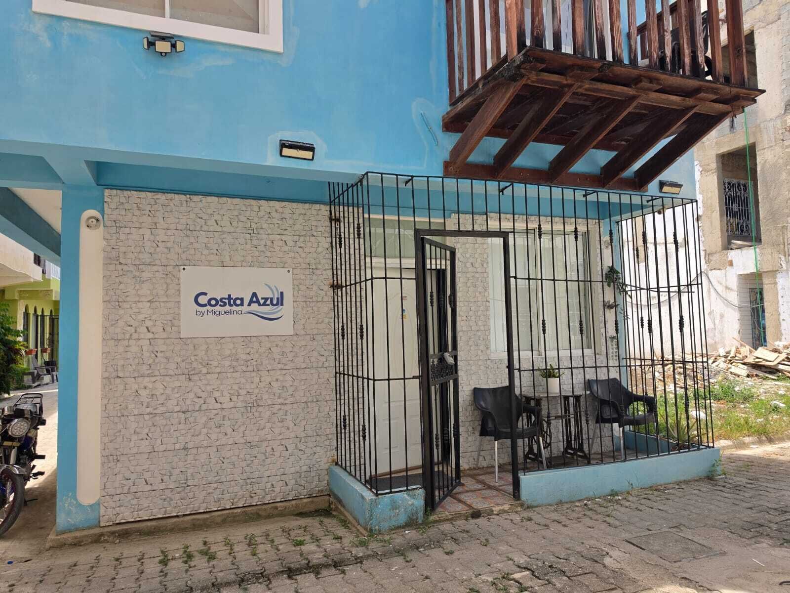 A building with a sign reading 'Costa Azul by Miguelina' and a gated patio area with outdoor seating.