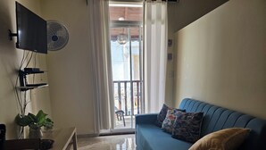 A living room with a blue sofa, a flat-screen TV, a ceiling fan, a potted plant, and a sliding glass door leading to an outdoor area. - Apartamento Costa Azul en El Pueblito (Puerto Plata)